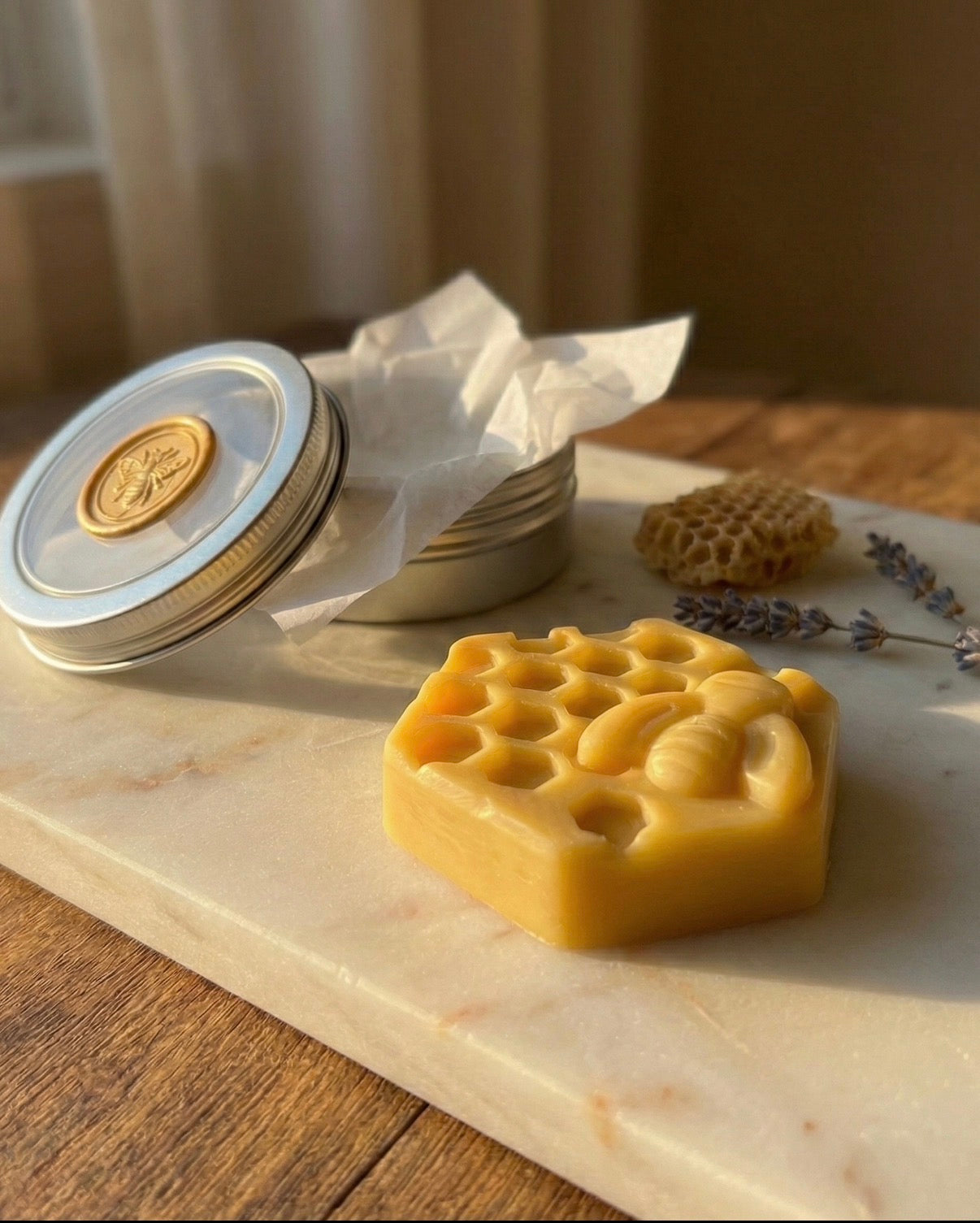 The Hive Bar packaging presentation. A silver reusable travel tin sits open on a marble surface, lined with white tissue paper and sealed with a gold wax seal stamp.